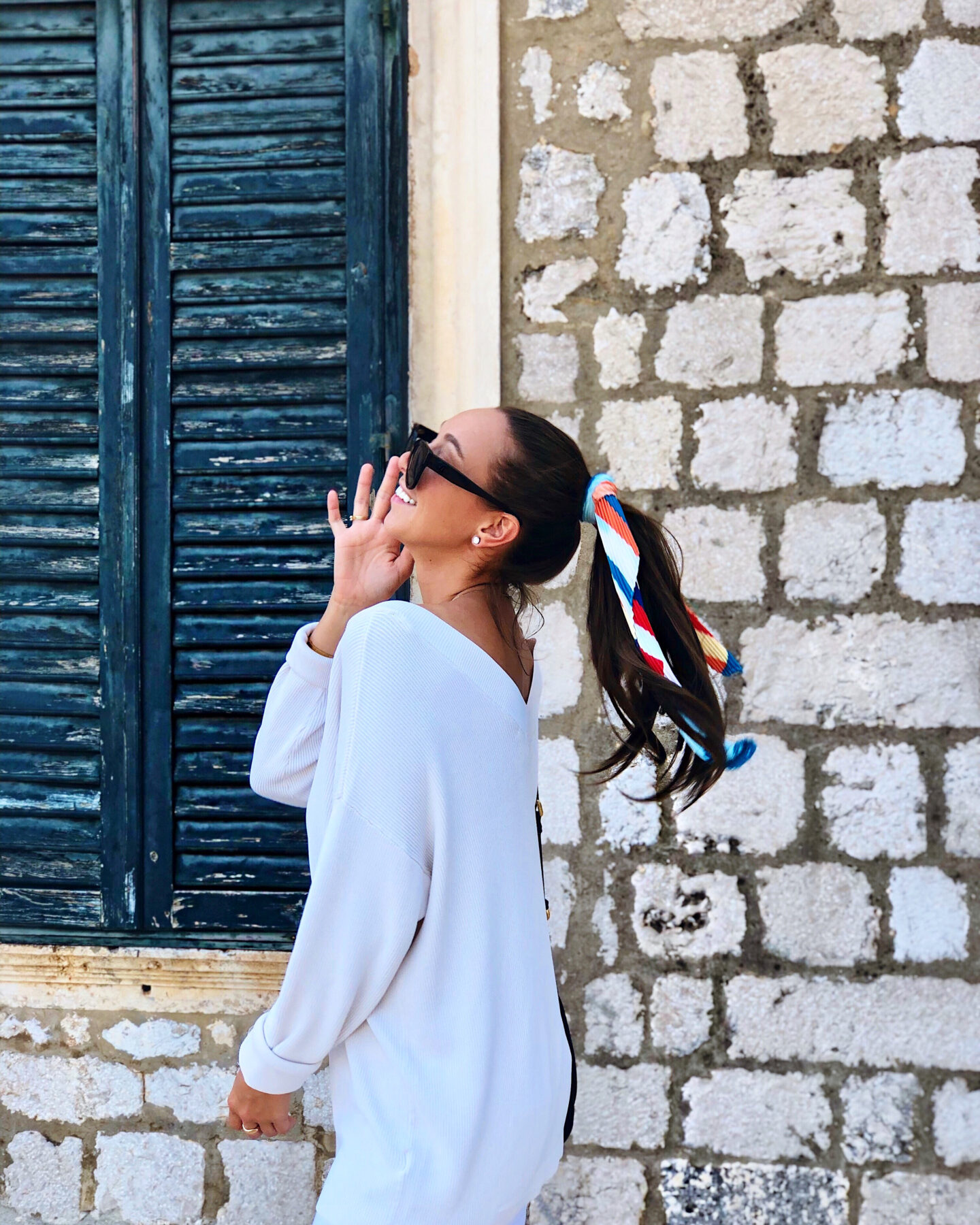 profile of woman wearing white sweater with hair in pony tail and scarf pushing up her sunglasses standing in front of stone wall and window with blue shutters in Dubrovnik Croatia