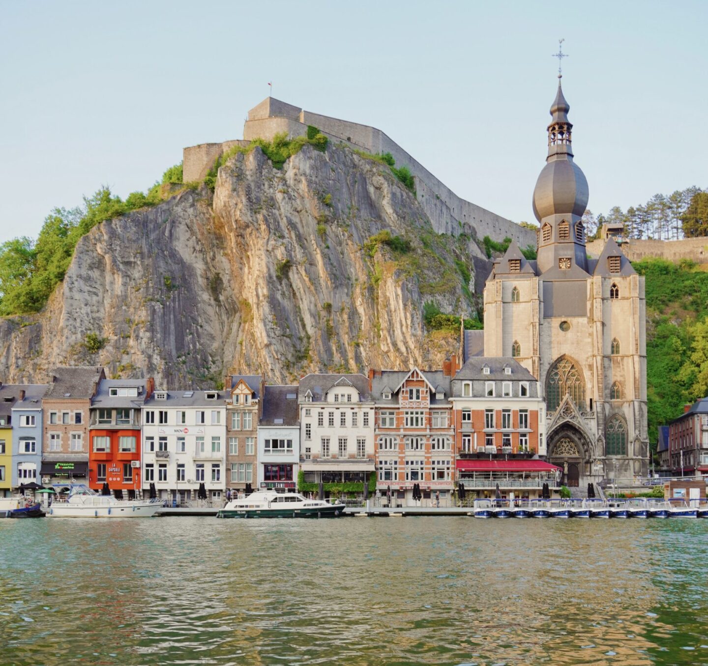View of Dinant city in Belgium with buildings lining the river and mountain in the background.