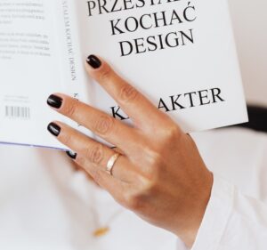 white woman's hand wearing a white shirt reading a white book  with finger nails painted with black nail polish 