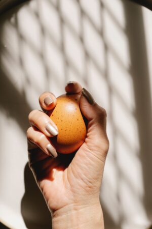 white woman's hand holding an orange with finger nails painted with brown chrome powder against a white background with a shadow.