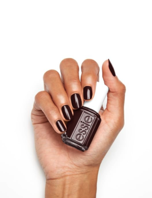 woman's hand holding an Essie nail polish bottle with finger nails painted with maroon nail polish against a white background.