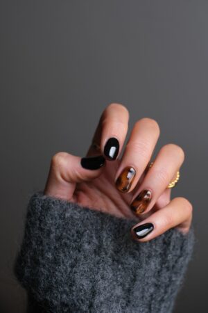 white woman's hand with finger nails painted black and brown with a tortoise shell design. wearing a grey sweater against a grey background.