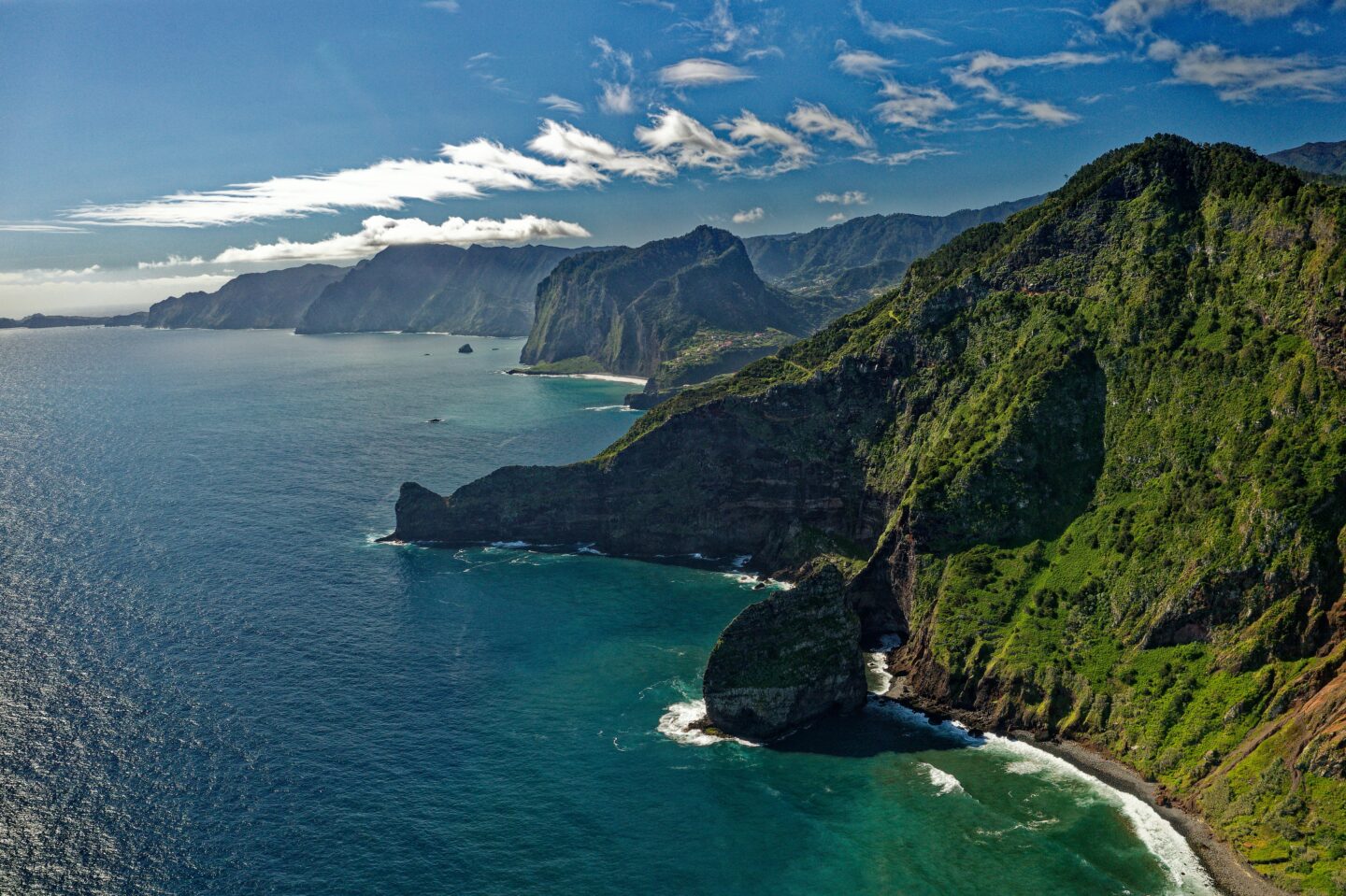 Ariel view of the coast of madeira showcasing the deep blue ocean meeting the dramatic, lush, green, mountain coastline with blue sky