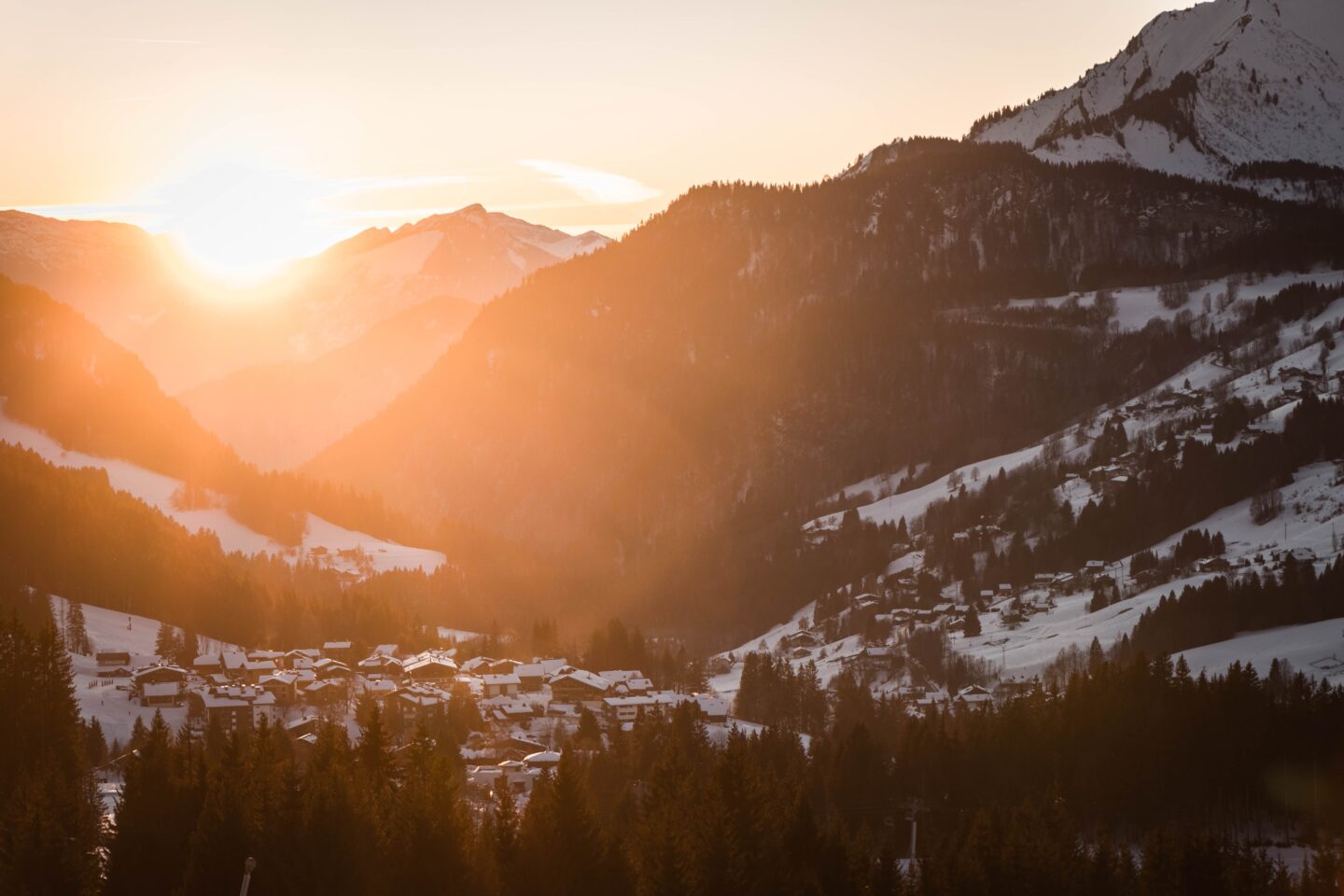 ski town in europe with orange glare of sun over mountain town in valley covered in snow
