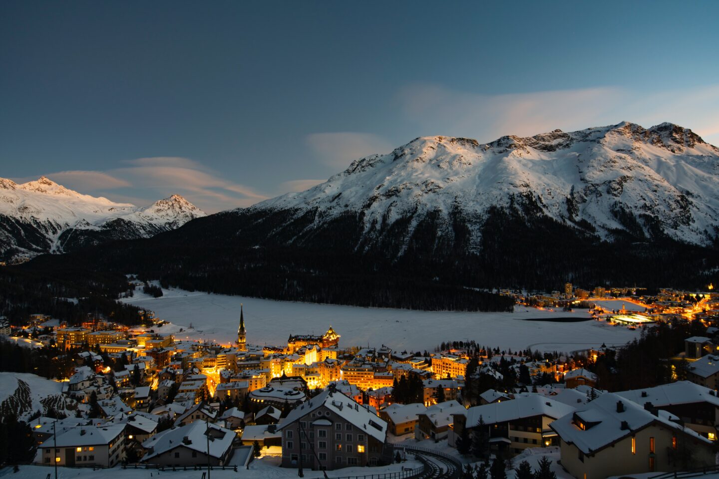 snow covered ski village in europe, chalets, lake and mountains at night, chalets with orange lights illuminated