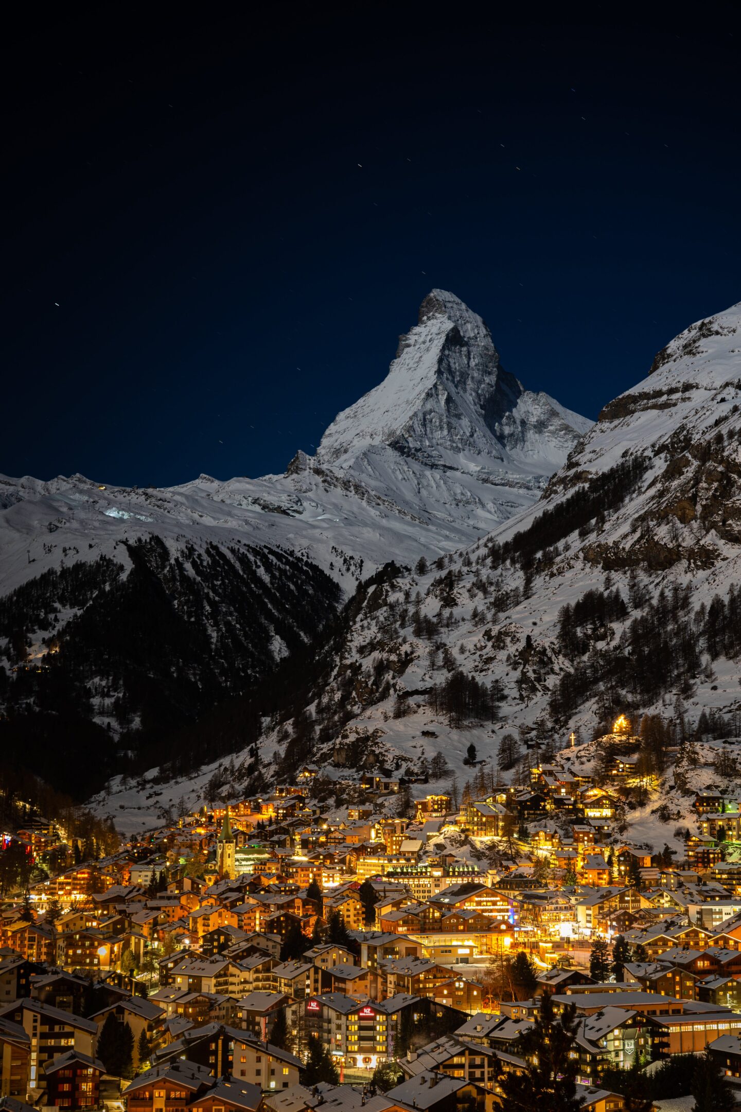 ski europe night in snow covered village with orange illuminated lights in valley between snow covered mountains and snow covered mountain peak in background with dark night sky 