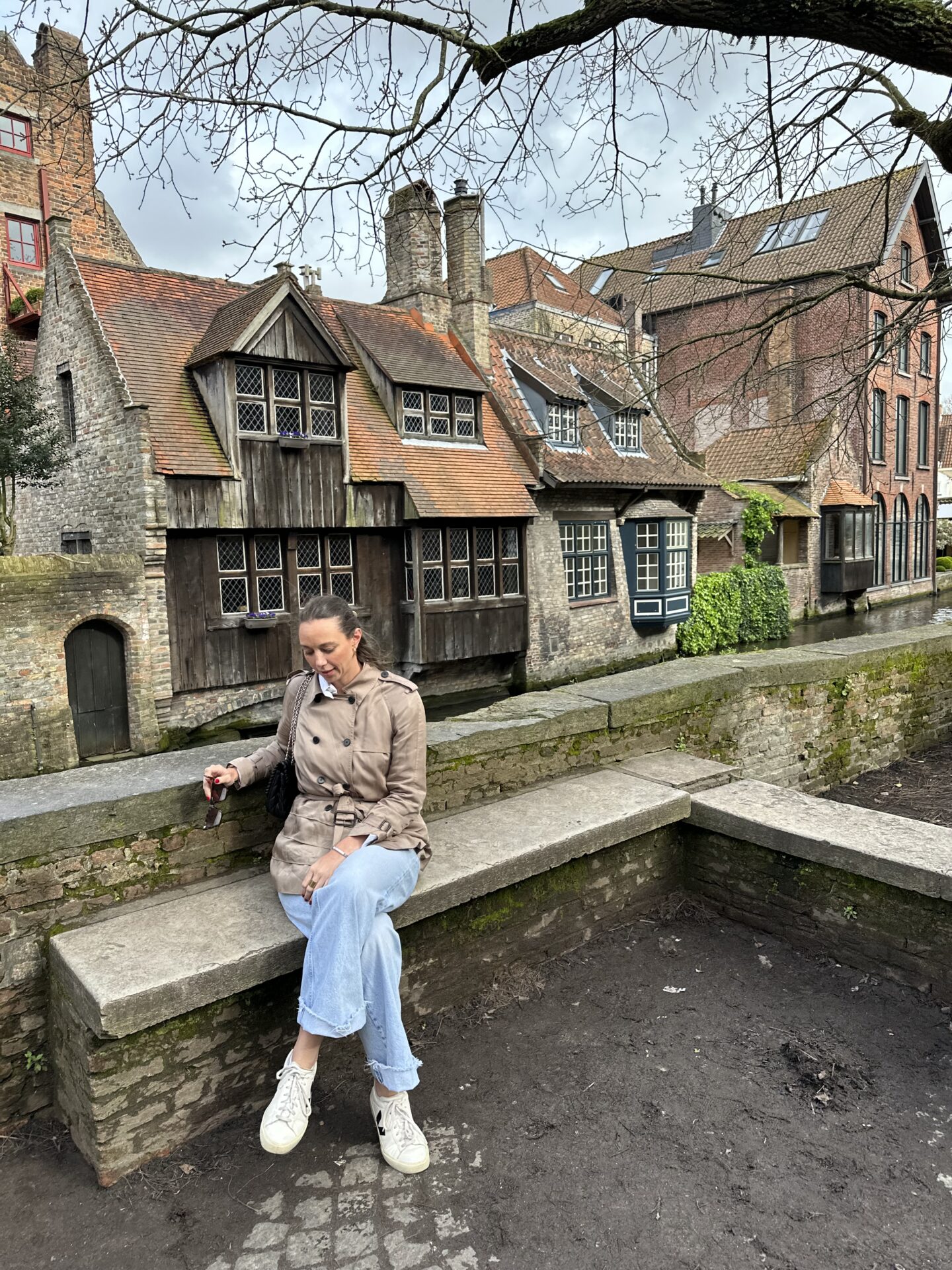 woman in brown trench coat and light blue jeans sitting on bench along river in bruges belgium with classic Belgian homes in background