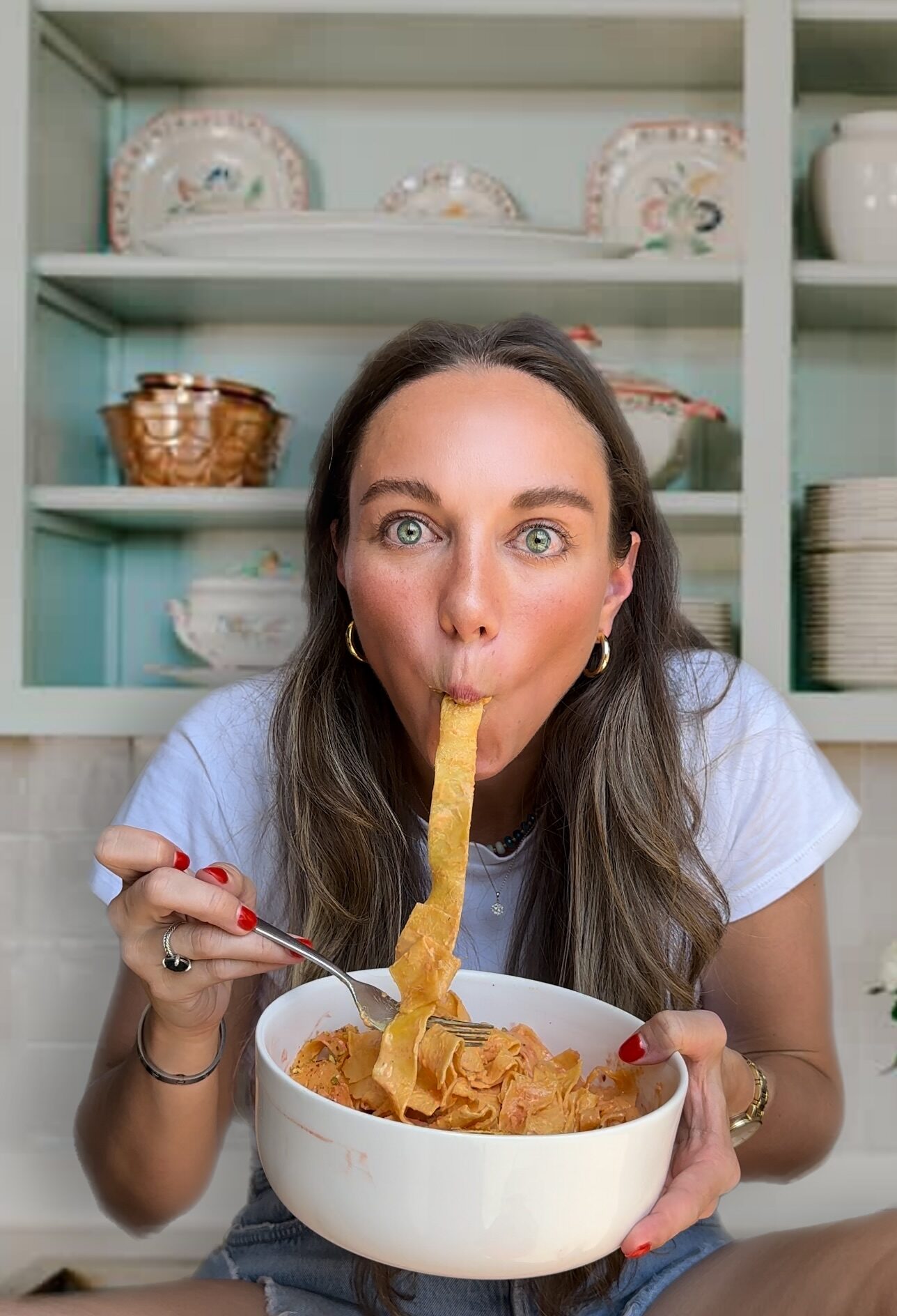 woman sitting in kitchen holding a bowl of bowl of pasta with a noodle in her mouth. Mikaela Higgins