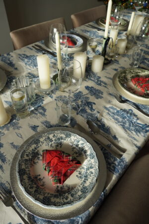 tablescape set for toile and tartan dinner party with toile de jouy tablecloth, blue, green and red flora dinner plates, tartan patterned napkins folded like Christmas trees and candles as the centerpiece