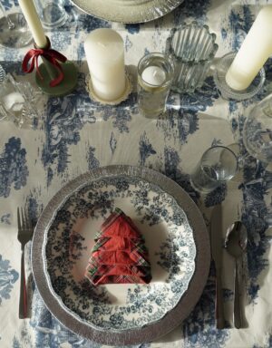 table place setting for toile and tartan dinner party with toile de jouy tablecloth, blue, green and red flora dinner plates, tartan patterned napkins folded like Christmas trees and candles as the centerpiece