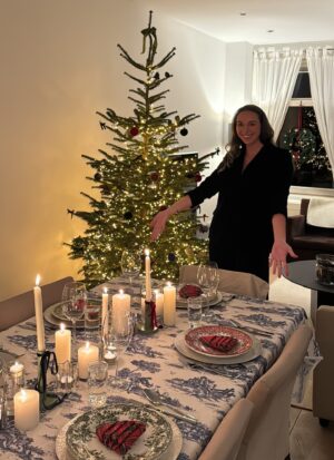 woman dressed festively standing by Christmas tree in her home displaying a set dinner table while hosting a toile and tartan holiday dinner party 